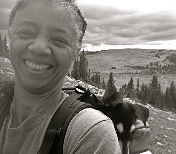 Black and white photo of Gillian Bowser on a mountain with her dog in her backpack peeking over her shoulder.