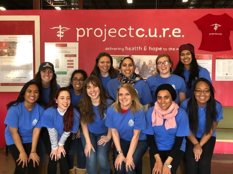 A group of Fulbrighters in blue shirts taking a posed photo in front of a red wall that says "project c.u.r.e."
