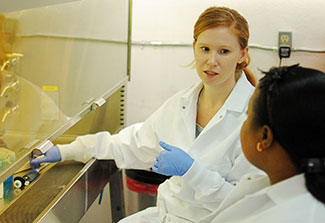 A woman in a lab coat with gloves on explaining something to another woman, also in a lab coat.