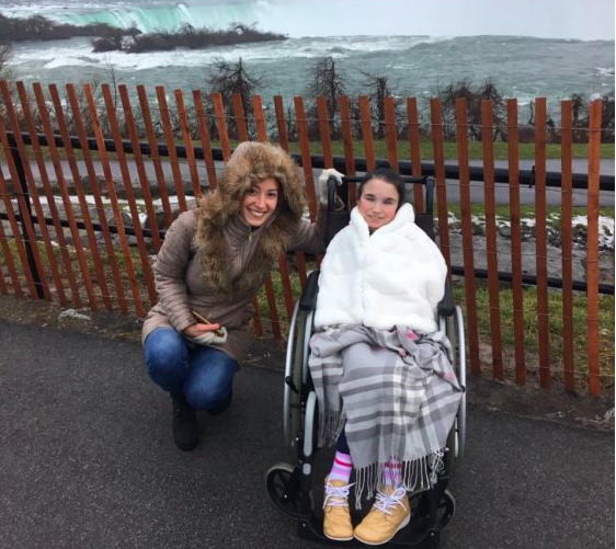 Two women posing in front of a waterfall. One is wrapped in two blankets and is on a wheelchair, the other kneels next to her in a hooded jacket. Both look cold, but are smiling.