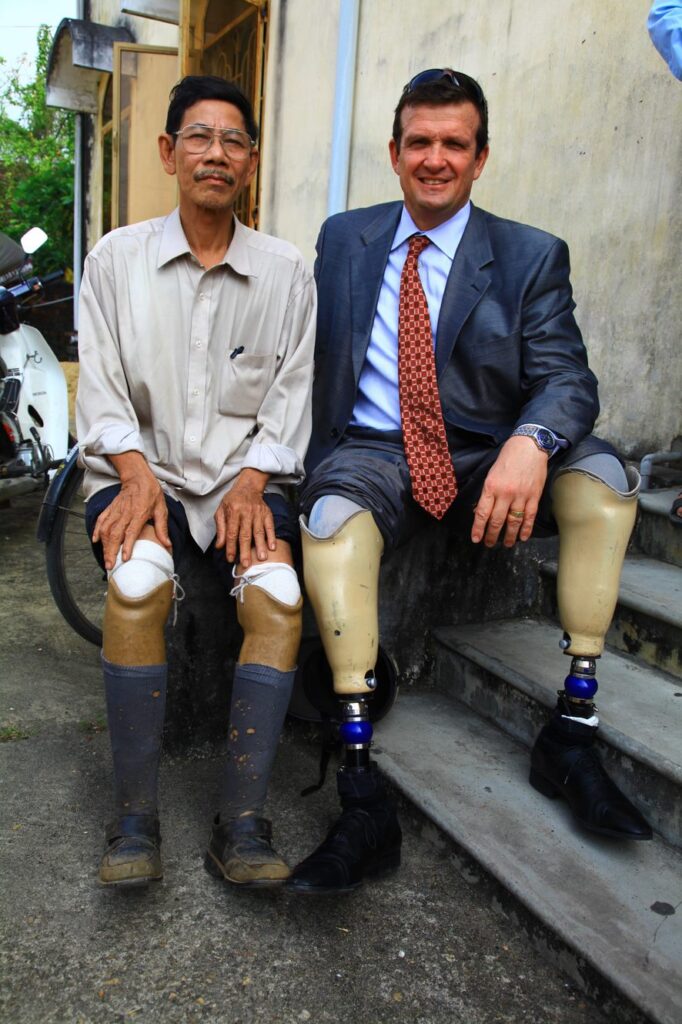 Ken Rutherford, in a suit with the legs rolled up to show his prosthetic legs, sitting next to a man in Vietnam who also has prosthetic legs.