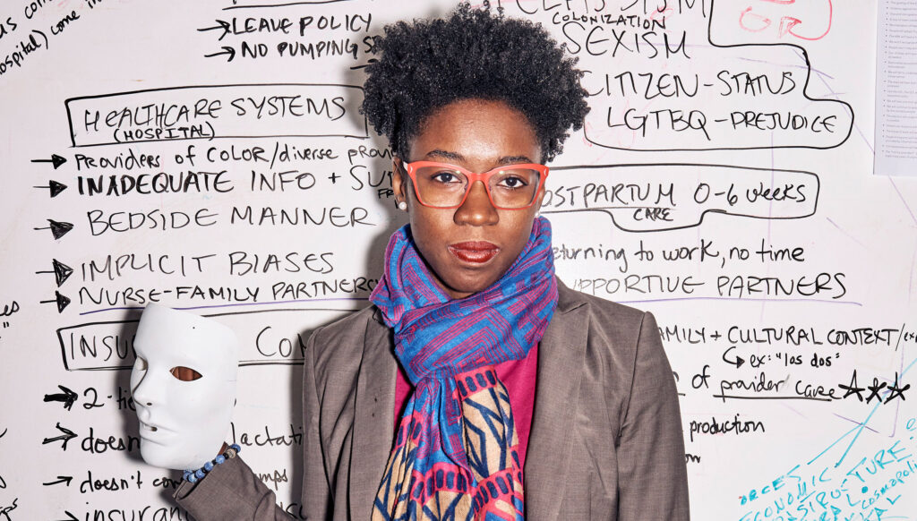 Joy Buolamwini in pink eyeglasses, a tweed blazer, and a a colorful scarf holding a white theater mask and standing in front of a whiteboard covered in phrases related to implicit bias and healthcare