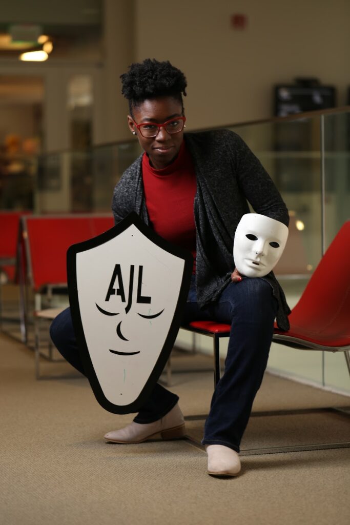 Joy Buowamlini sitting on a red chair with a red top and dark gray blazer holding a cardboard shield with the letters "AJL" and a smiley face with closed eyes in one hand and a white theater mask in the other.
