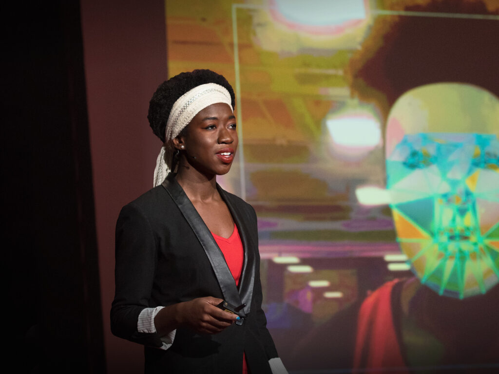  Joy Buolamwini wearing a red shirt, black blazer, and white wraparound headband giving her TED Talk on algorithmic bias. Photo Credit: TED Talk. 
