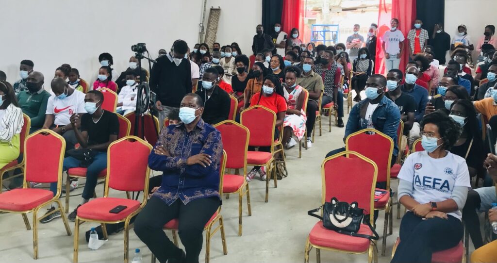 A room full of people, most wearing masks, sitting indoors on red chairs at a community event