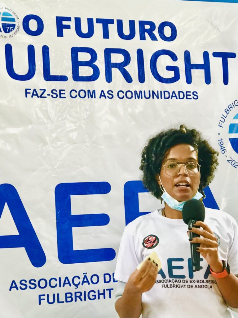 A woman with glasses and a mask pulled down on her face speaks into a mic she is holding in front of a banner that says "Fulbright AEFA"
