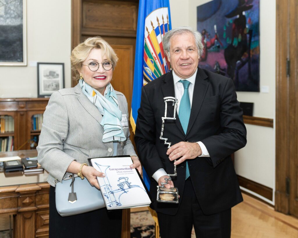 Hilda Ochoa-Brillembourg presents Luis Almagro (right) of the Organization of American States (OAS) with the 2018 Global Leadership Award on behalf of the Youth Orchestra of the Americas (YOA). Hilda wears a light gray blazer and light blue silk scarf and round glasses and holds a light blue bag and a set of thin books in both hands. Luis wears a black suit with an aqua tie, holding a trophy.