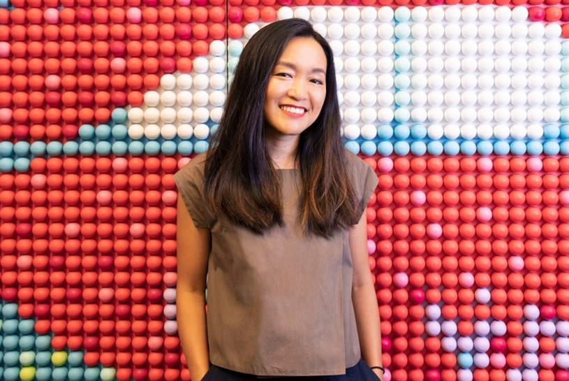 Soyoung Kang with a casual top standing in front of a wall made of small balls in different colors - mostly red, pink, white, and blue