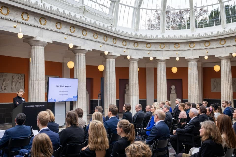 Photo of an audience from the side, viewing a presentation in a fancy building with pillars and transparent roof.