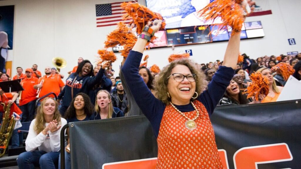 Photo of a college sports game. Students sit in the bleachers wearing orange and navy blue, the school's colors, cheering on the team. A woman is in the front, the center of the photo, wearing orange and navy blue, and waving similarly colored pompoms.