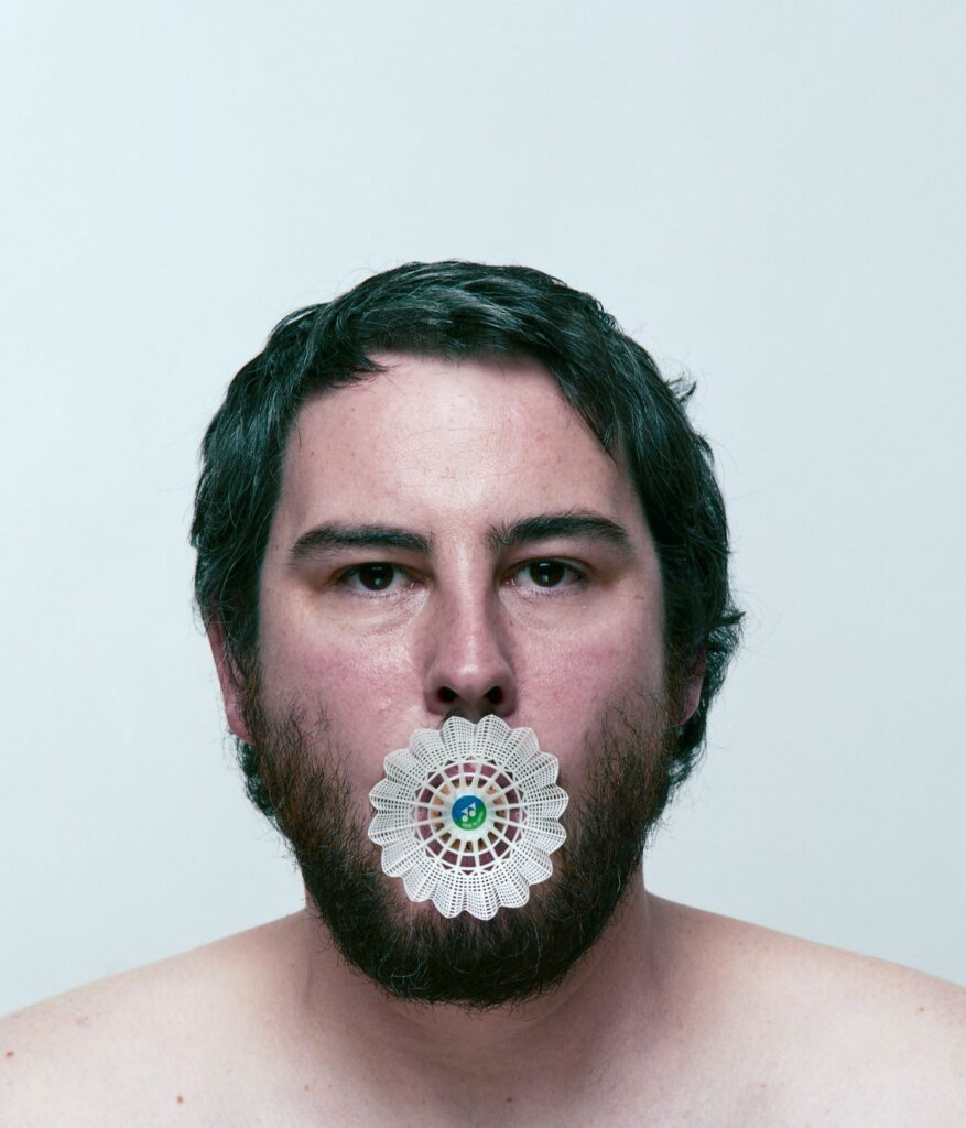 Artistic photograph of a person with dark hair and a dark beard with a daisy-type flower in their mouth against a pale blue background.