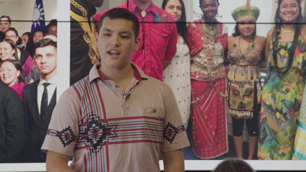 Person wearing a tan polo with a design standing in front of a backdrop of people dressed in traditional cultural clothing