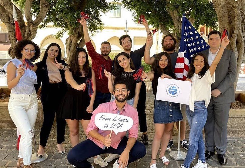 Group of people posing outside, one holding a sign that says "From Lebanon to USA" and another holding the Fulbright logo. Someone in the back is holding the US flag.