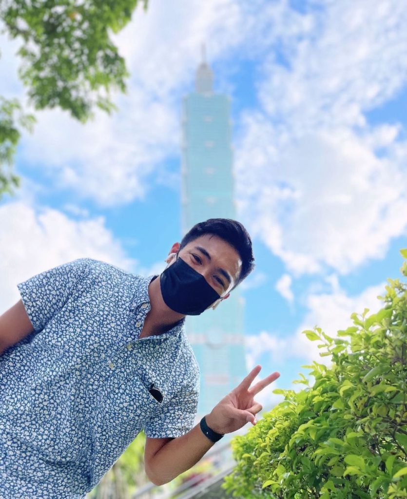 Person with short black hair and blue button-down and a dark mask posing with a peace sign outdoors