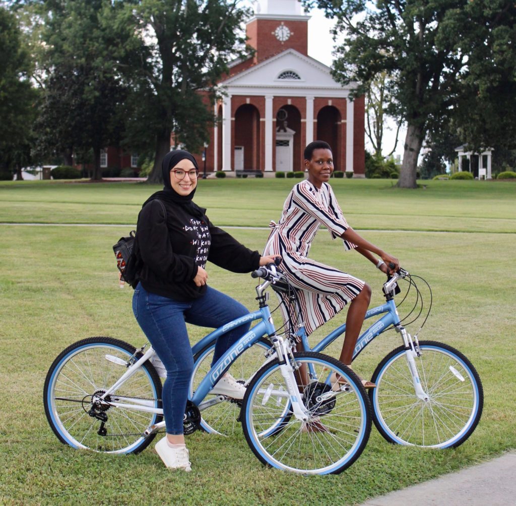 Two people outdoors posing on bikes. One is wearing a striped dress with very close-cropped black hair, the other is wearing jeans and a sweatshirt with a black hijab