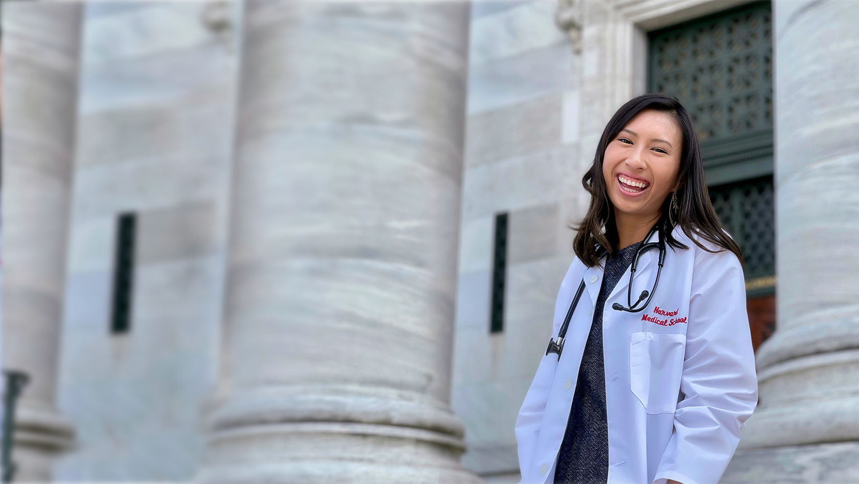 Person in lab coat and stethoscope laughing on steps of building with pillar in background